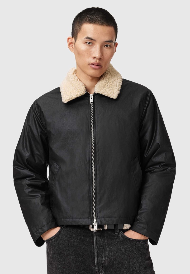 Chaqueta de cuero negra con un collar de borrego beige, cierre de cremallera, bolsillos laterales y un acabado texturizado. El diseño es contemporáneo y ajustado.