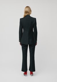 Woman with long blonde hair wears a tailored black suit, red socks, and white high-heeled shoes, standing facing away on a plain background.