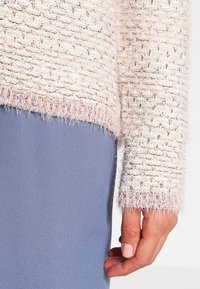 Primer plano de una mano descansando al lado del cuerpo, vistiendo un suéter de punto texturizado en color beige-rosado y una falda o pantalones azules lisos.