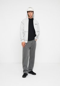 Lys grå hettegenser med svarte detaljer og logo, kombinert med en svart t-skjorte og løse grå jeans. Svarte joggesko fullfører antrekket.