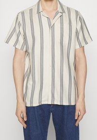 Camisa de manga corta en tela beige claro con rayas verticales grises, cuello con muesca, bolsillo frontal y cierre con botones.