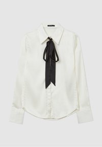 Blouse blanche à manches longues avec grand col et nœud papillon en satin noir au niveau du cou, posée à plat sur un fond clair.