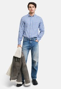 Blå og hvit rutete skjorte med knapper, lyseblå jeans, grå jakke med pelsfôr over armen, svarte støvler.