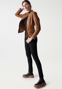 Chaqueta de cuero marrón con forro de contraste, diseño ajustado, bolsillos con cremallera, combinada con jeans skinny negros y botas de tobillo negras de suela gruesa.