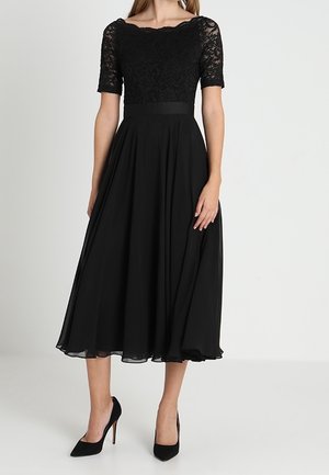 Femme porte une robe midi noire avec des manches courtes en dentelle et une taille cintrée, associée à des chaussures à talons hauts noires sur un fond uni.