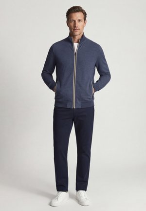 Man draagt een marineblauwe jas met rits, donkere broek en witte sneakers, staat met handen in de jaszakken tegen een effen achtergrond.