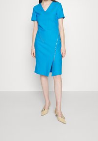 Ljust blå klänning med korta ärmar, V-hals, frontdragkedja och knälång fåll. Kombinerad med beige klackar med en böjd design.