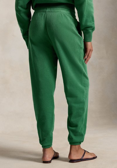 Osoba ubrana w zielone spodnie dresowe i pasującą bluzę, stojąca boso, obok ciemnych klapek, na neutralnym tle podłogi.