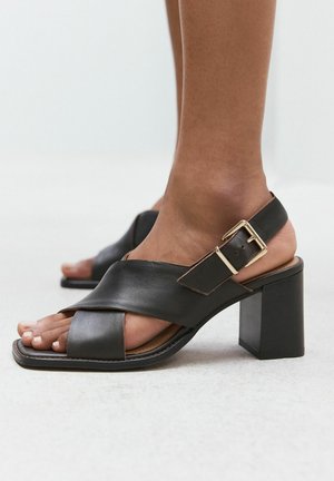 Zwarte leren sandalen met hak, brede gekruiste bandjes en een gouden gesp op een donkerhuidige voet tegen een neutrale achtergrond.