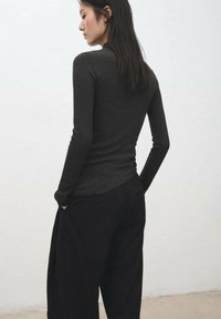 Black ribbed long-sleeve top with a fitted design, paired with loose black trousers. The model has hands in pockets, showcasing the outfit's silhouette.