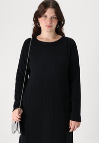 Abito nero a maniche lunghe in maglia testurizzata, con cuciture verticali, scollatura rotonda e una borsa a tracolla in metallo con finitura metallica.