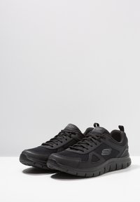 Black athletic shoes with a mesh upper, textured accents, and a cushioned sole. Features laces and a pull tab at the back.