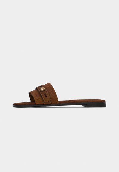Sandale mule en suède marron avec une semelle noire plate et une large bride ornée d'une petite boucle décorative et d'un détail en forme de trou de serrure.