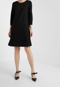 Musta polvipituinen mekko, jossa on kolmennesosa hihasta, valmistettu sileästä kankaasta. Mekossa on istuva yläosa ja liehuva hame; sitä on yhdistetty mustiin kärkitossuihin.
