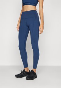 Høytlivsnavy leggings laget av elastisk materiale, med sidelomme, kontrasterende sømmer og en tettsittende design. Svarte treningssko.