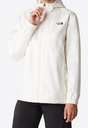 Persona con una chaqueta blanca impermeable North Face con capucha, cerrada con cremallera, y pantalones negros sobre un fondo liso.