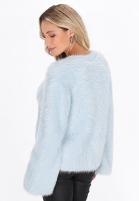 Frau mit langen blonden Haaren, die silberne Creolen und einen hellblauen, flauschigen Pullover trägt und seitlich vor einem weißen Hintergrund posiert.