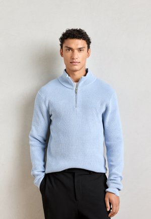 Light blue knit pullover with a half-zip neck, ribbed texture, and long sleeves; paired with black trousers for a clean, modern look.