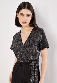 Jonge vrouw met donker haar draagt een zwarte sequined wraptop met korte mouwen en een gestrikte taille tegen een lichte effen achtergrond.