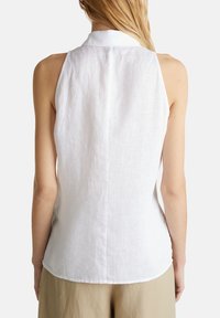 Femme portant un haut blanc sans manches avec col et un pantalon beige, vue de dos sur un fond uni.