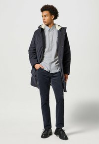 Parka azul marino con un interior texturizado y suave, combinada con una camisa abotonada de color gris claro y pantalones de pana oscuros; zapatos negros de cordones.