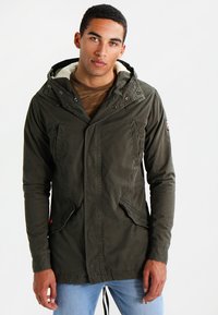 Giovane uomo che indossa una giacca con cappuccio verde scuro foderata in pile, sopra una camicia marrone e jeans azzurri chiari, in piedi davanti a uno sfondo bianco.