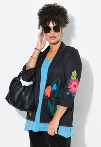 Schwarzes Blazer mit buntem Blumenstickerei an den Ärmeln, kombiniert mit einem blauen Oberteil. Das Modell hält eine schwarze Handtasche und trägt übergroße Sonnenbrillen.