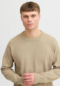Beige knitted sweater with a round neckline, featuring a textured fabric and ribbed cuffs, worn by a model with short hair and a neutral expression.