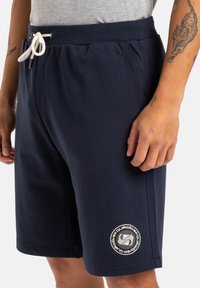 Pantalones cortos azul marino hechos de una tela suave con cintura ajustable mediante cordón, con un parche de logo circular en el muslo izquierdo.