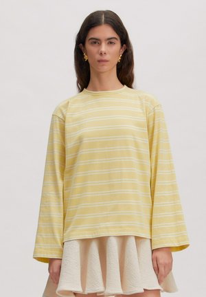 Vrouw met lang donker haar draagt gouden abstracte oorbellen, een geel-wit gestreept shirt met lange mouwen en een getextureerde crèmekleurige uitlopende rok.