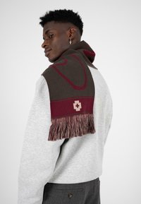 Strikket tørklæde i mørkebrun og maroon med en struktureret finish, prydet med frynser og et geometrisk mønster, suppleret med et logo.