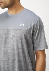 Szürke sportpóló, texturált mintával és sima ujjakra. Az elején fehér Under Armour logó található. Rövid ujjú kivitel.