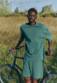 Jeune homme en tenue verte se tenant dans un champ d'herbe, tenant un vélo noir, avec des cheminées industrielles visibles à l'arrière-plan.