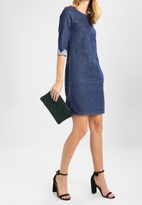 Vestido vaquero con mangas tres cuartos, con escote redondo y bolsillos delanteros. Combinado con tacones negros de tiras y un bolso de mano verde.