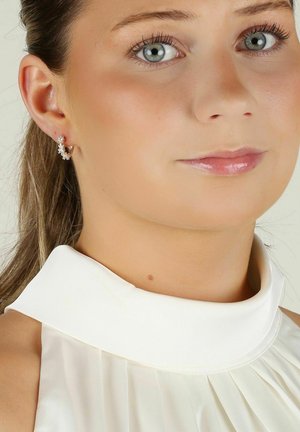 Silverfärgade hoops med blomaccent inlagda med små klara stenar. Modellen bär en vit, plisserad krag topp. Neutralt bakgrund.