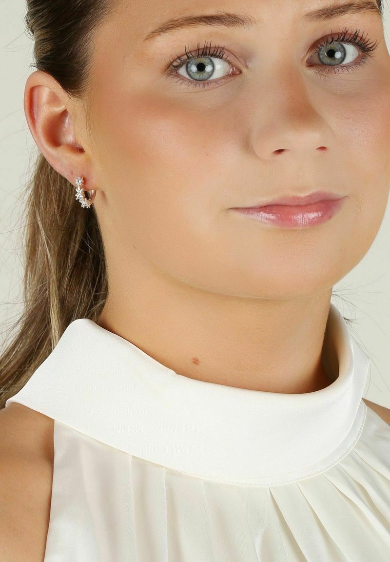 Silverhoopsörhängen med blomsterdetaljer, genomskinliga ädelstenar och en slät vit högkragad topp med veck, mot en neutral bakgrund.