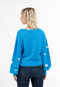 Maglione azzurro brillante e soffice con maniche larghe, dotato di colorati patch a forma di cuore sulle maniche. Abbinato a jeans scuri.
