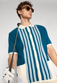 Teal og krem kortermet golf polo med vertikale striper, knappelukking og sidelommer. Materialet ser teksturert ut, med krage detaljer.