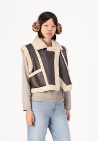 Brun lædervest med beige shearlingkant. Har en høj krave og åben frontdesign. Båret over en lys grå sweater og blå jeans.