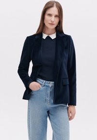 Navy fluwelen blazer met een gekartelde revers, gedragen over een geribbeld donker sweater en lichte, hoge taille jeans. De blazer heeft één zak.