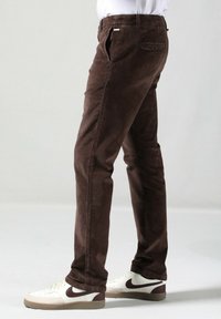 Hnědé kordové kalhoty slim fit s hladkým povrchem, bočními kapsami a poutky na opasek, doplněné bílými teniskami.