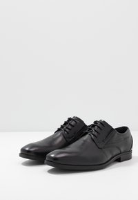 Chaussures de ville en cuir noir avec des orteils arrondis, un laçage classique, des accents de couture subtils et une finition lisse et brillante. Semelle en caoutchouc.
