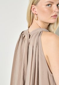 Mujer rubia con top beige sin mangas y espalda plisada, y pendiente de aro dorado retorcido, mirando por encima del hombro.