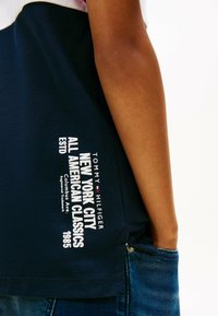 Marineblå polo T-shirt med logo og tekstdetaljer i hvid, med et struktureret stof og en slidssplit. Synlige sømme i kanten.
