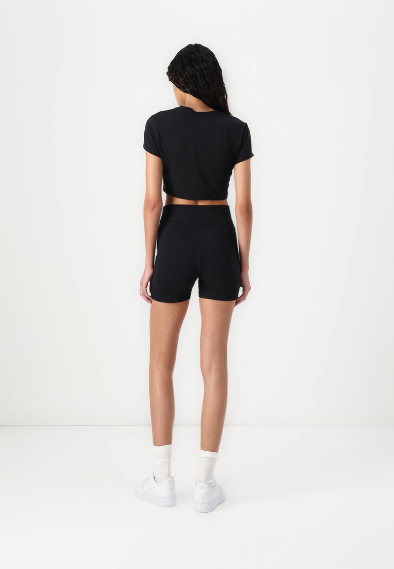 T-shirt noire courte associée à un short noir taille haute. Le matériau a l'air lisse ; le short est ajusté. Baskets blanches avec des chaussettes blanches.