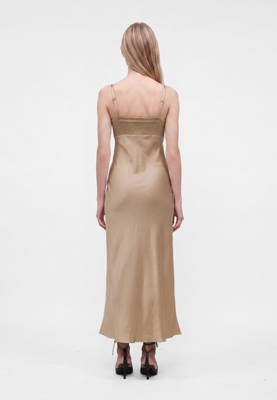 Femme aux longs cheveux blonds, vêtue d'une robe nuisette en satin doré et de chaussures à talons noires, debout de dos sur un fond blanc.
