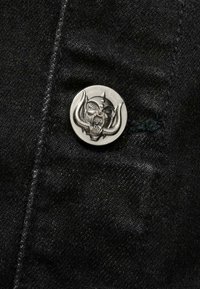 Svart denimtyg med en silverfärgad knapp som har en detaljerad design av ett kranium med horn. Texturen är lätt grov och framhäver materialet.