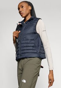 Colete acolchoado azul-marinho com textura matelassada, gola alta e bolsos com fecho de correr, combinado com uma blusa de manga longa leve e calças verde-oliva.