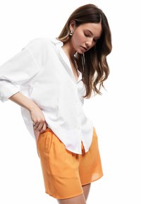Camicia bianca a bottone con maniche arrotolate, abbinata a pantaloni corti arancioni e texture. Il modello è posato in modo elegante, mostrando i dettagli del capo.