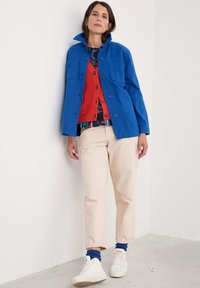 Blue jacket with collar and buttons, red cardigan, patterned shirt, cream pants, white sneakers, blue socks, standing against a plain background.